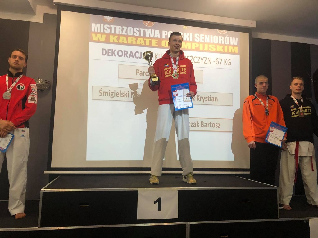 Michał Parczewski na najwyższym podium, trzymający puchar i dyplom za zajęcie 1 miejsca. W tle napis: Mistrzostwa Polski Seniorów w karate olimpijskim. Na podium widoczni pozostali zawodnicy.